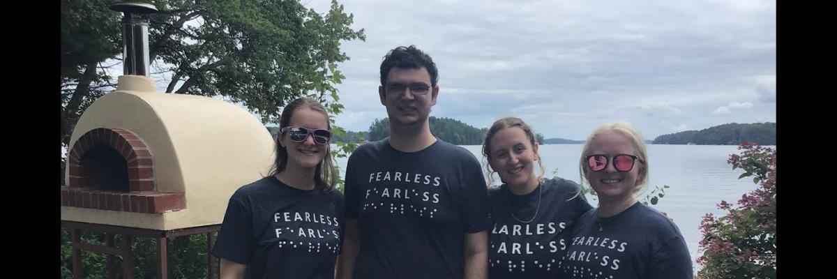 From left to right: (L-R) Emilee Schevers, Tavish Gillan, Taylor Gaudon and Jade Ondrik by the outdoor pizza oven at CNIB Lake Joe. They are wearing blue t-shirts with the word “Fearless” in print and braille.