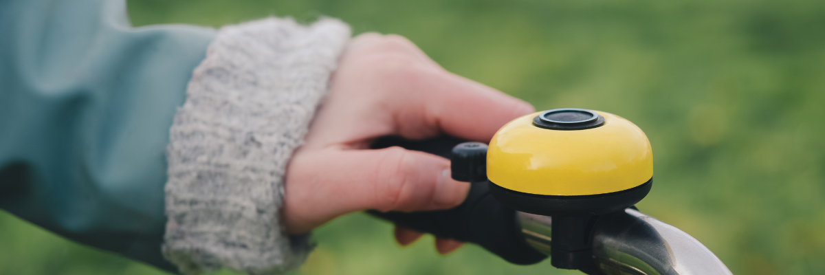 A person’s hand rings a yellow bicycle bell. The bell is mounted on the bicycle handlebar. 
