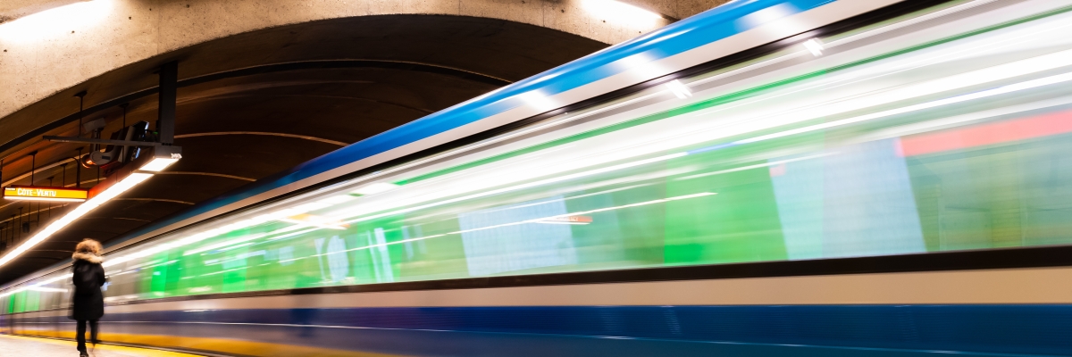 A train pulls into an empty indoor station. The image is out of focus and blurry, suggesting that the train is in motion as it approaches its stop. The station has a curved ceiling with lights, and one person walks along the platform.