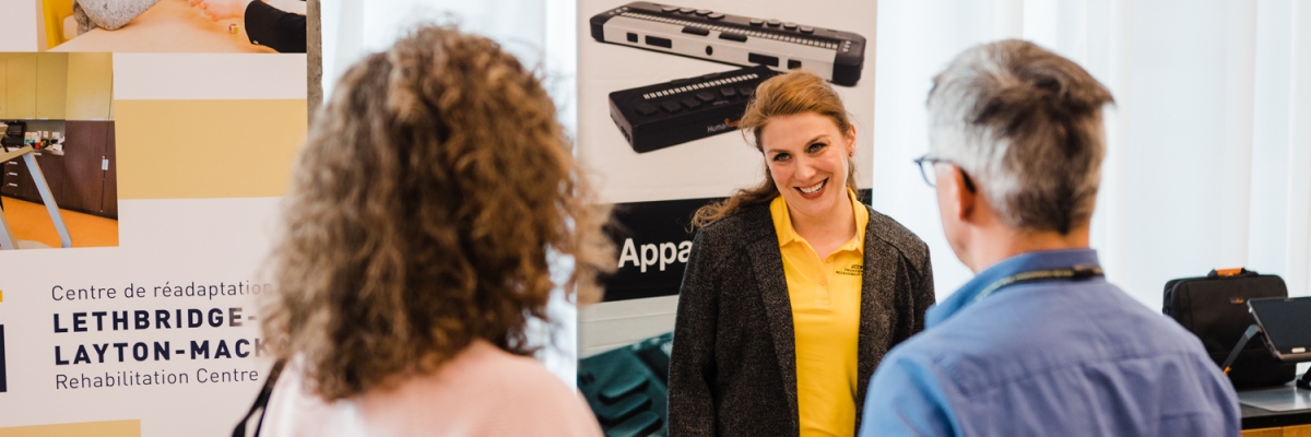 A woman behind an exhibit table speaking to a man and woman.