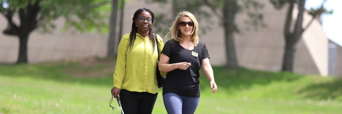 A woman provides sighted guide to another woman who is navigating with a white cane.