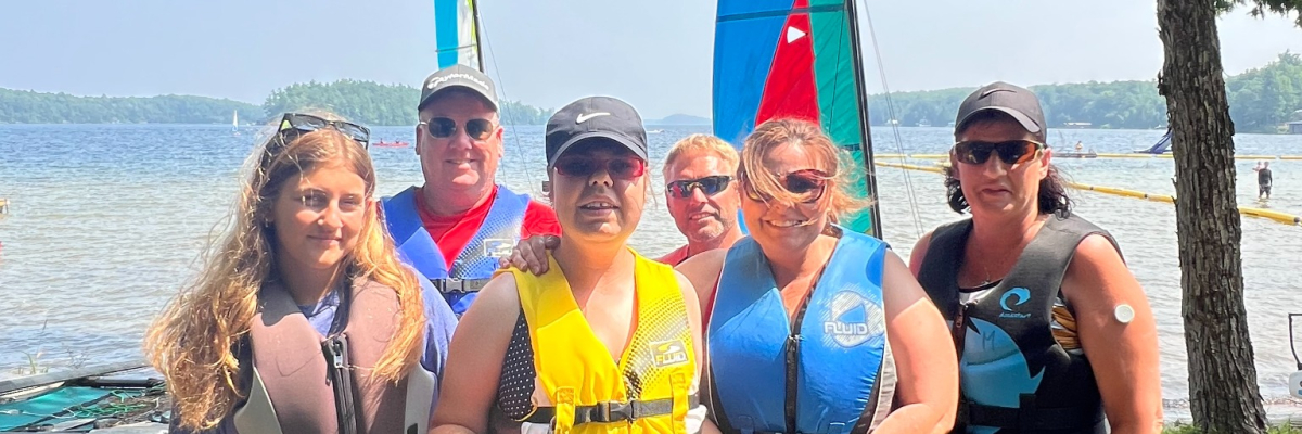 Six people are standing in front of the lake with sailboats them. They are all wearing life jackets and smiling at the camera