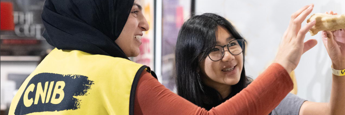 Two women, one wearing a CNIB vest, smile while holding and examining an object together indoors.