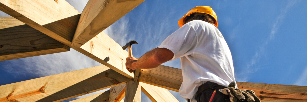 Construction worker wearing a hard hat uses a hammer to secure wooden roof beams, viewed from below against a bright blue sky.