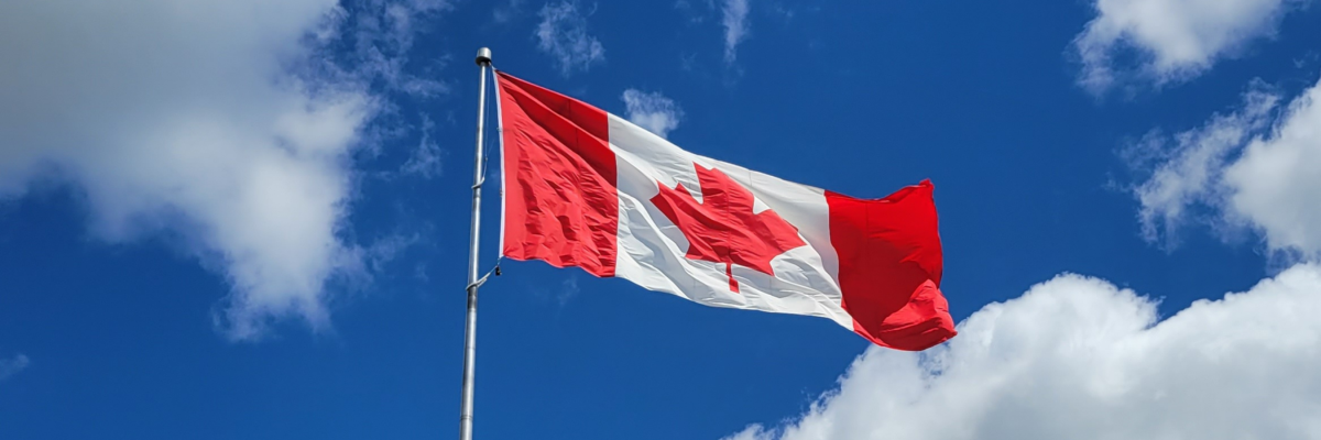 A Canadian flag waves on a flagpole against a bright blue cloudy sky.