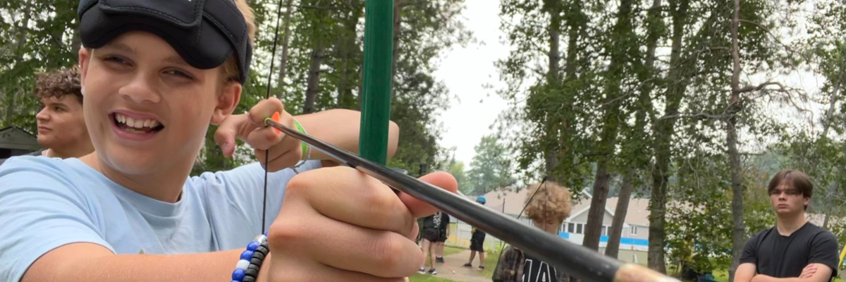Young camper with blindfold is drawing a bow and arrow.
