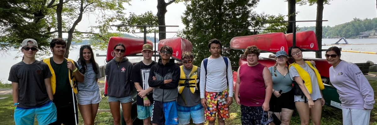 A group of youth are standing outside in front of stacked canoes. 