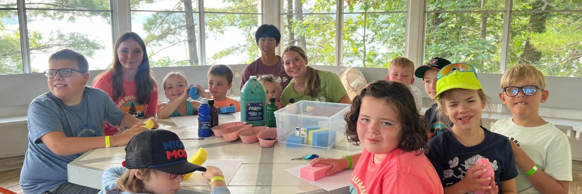 Children sitting in a gazebo at a wooden table covered with craft supplies.