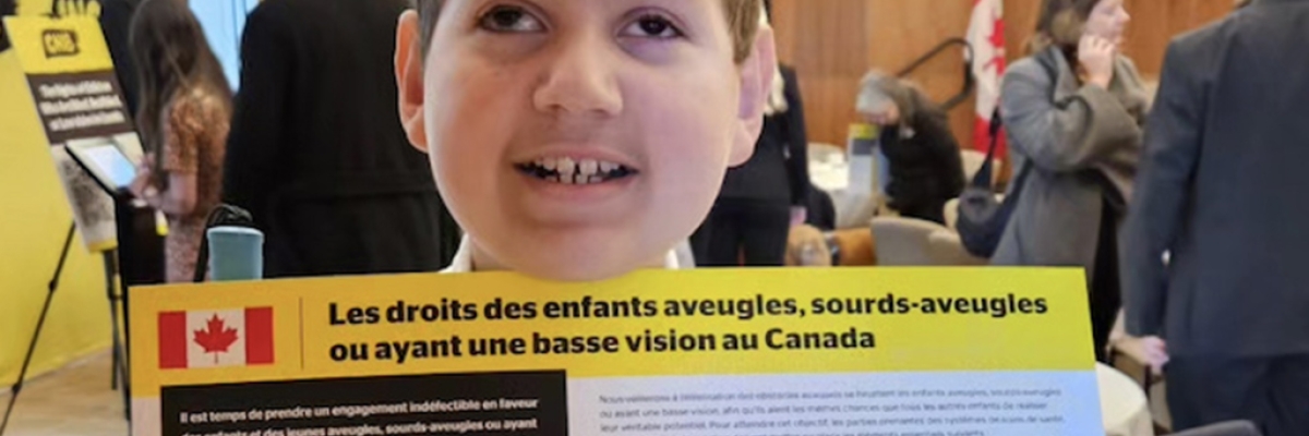 A boy is standing in a meeting room with business people behind him. He is holding up the Rights of Children Who Are blind in Canada (French version).