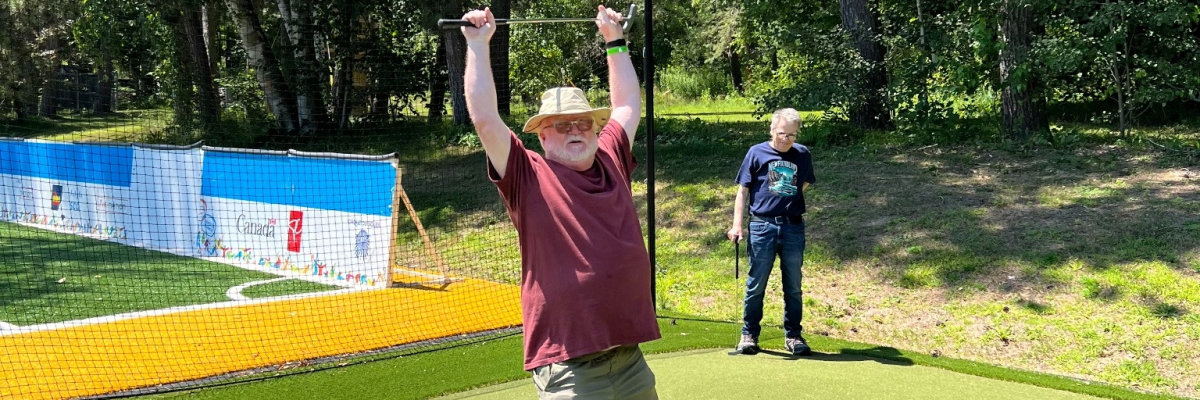 A man is standing in a mini putt course triumphantly holding a club over his head and smiling.