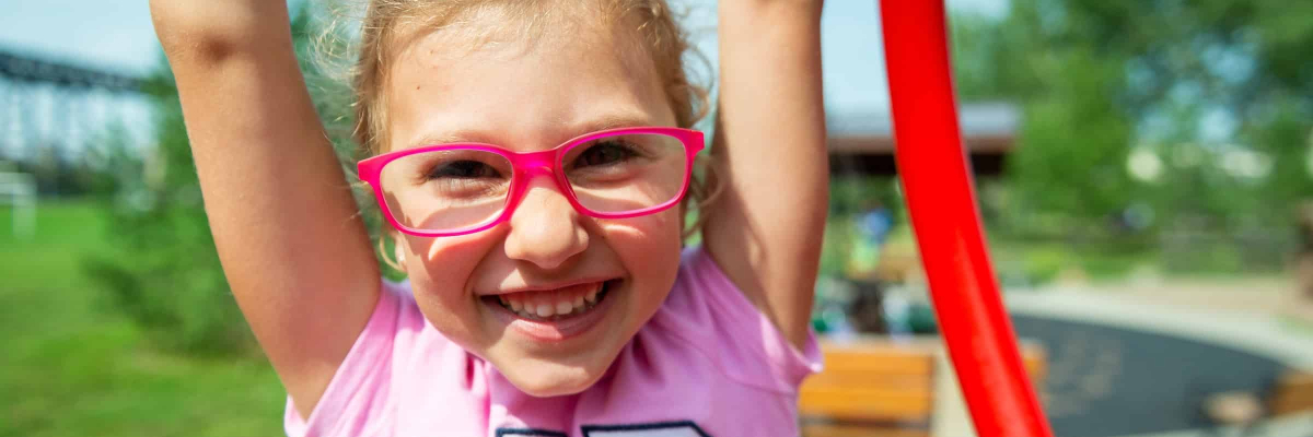 Girl wearing red glasses and pink shirt is smiling and holding her arms over her head.