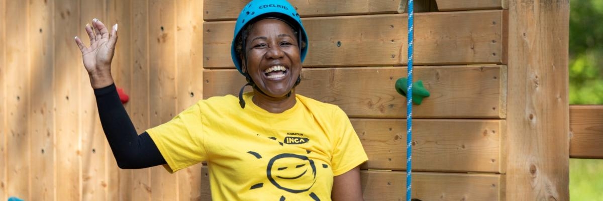 A black woman is standing in front of the climbing tower, smiling and waving with one hand. She is wearing a blue helmet and a yellow CNIB tshirt with a smiley face.
