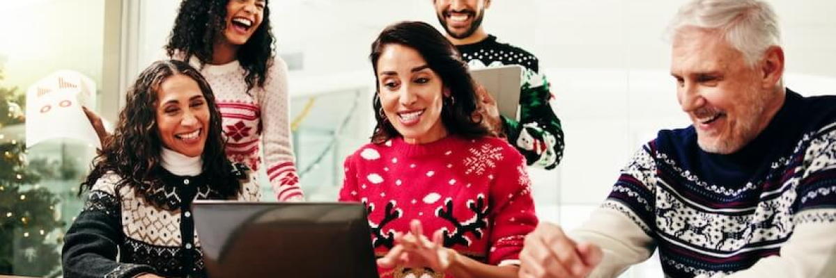Four men and women wearing holiday sweaters and Santa hats sit around a laptop.