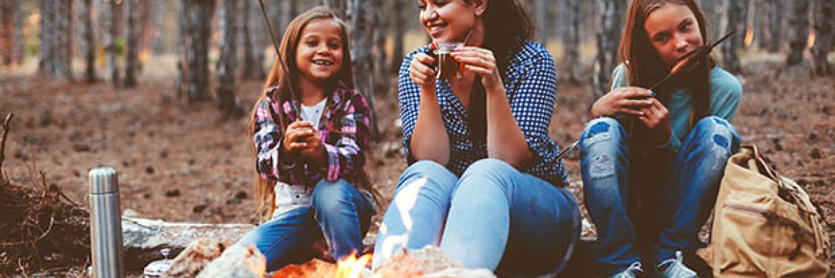 A woman and two girls sit outside in front of a fire eating treats.