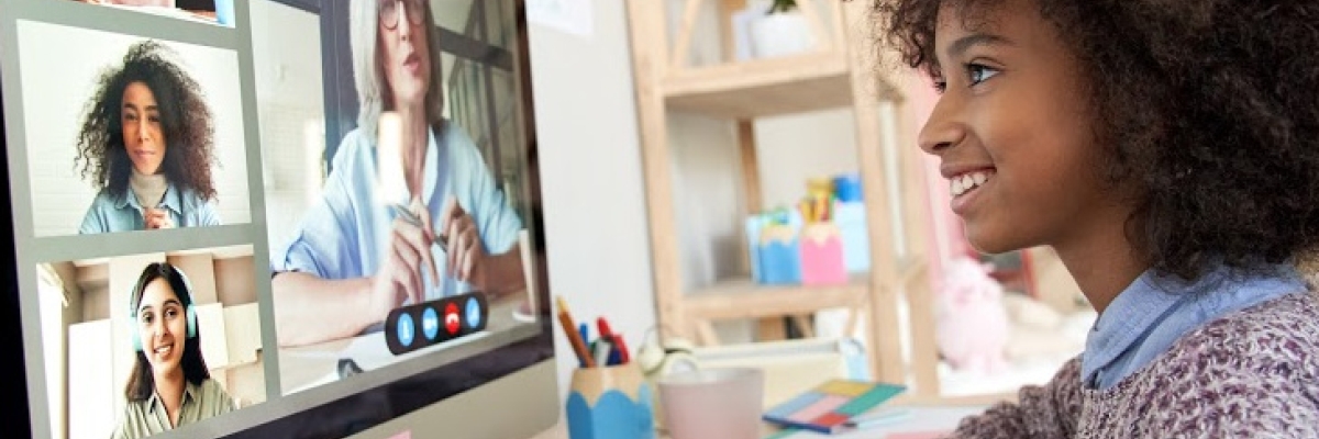 A young girl is sitting at a desk, looking at a computer screen with other people on screen.