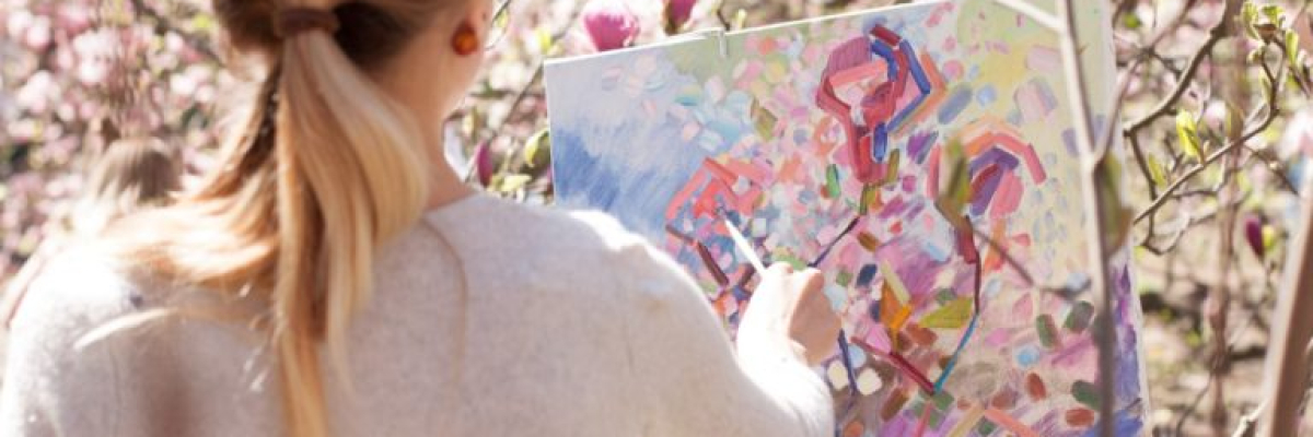 A woman is standing outside amongst bushes with blossoms. She is painting on a canvas.