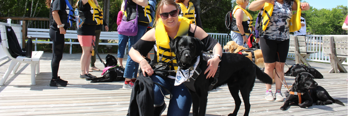 A woman is kneeling on a dock with her arm around a black lab. There are people behind her with dogs. 