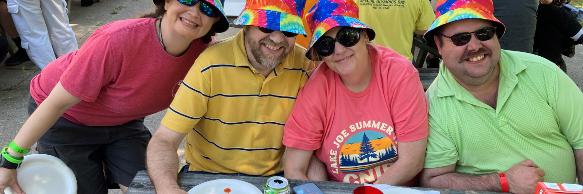 Four adults sitting outside at a picnic table wearing colourful tshirts and tye die bucket hats