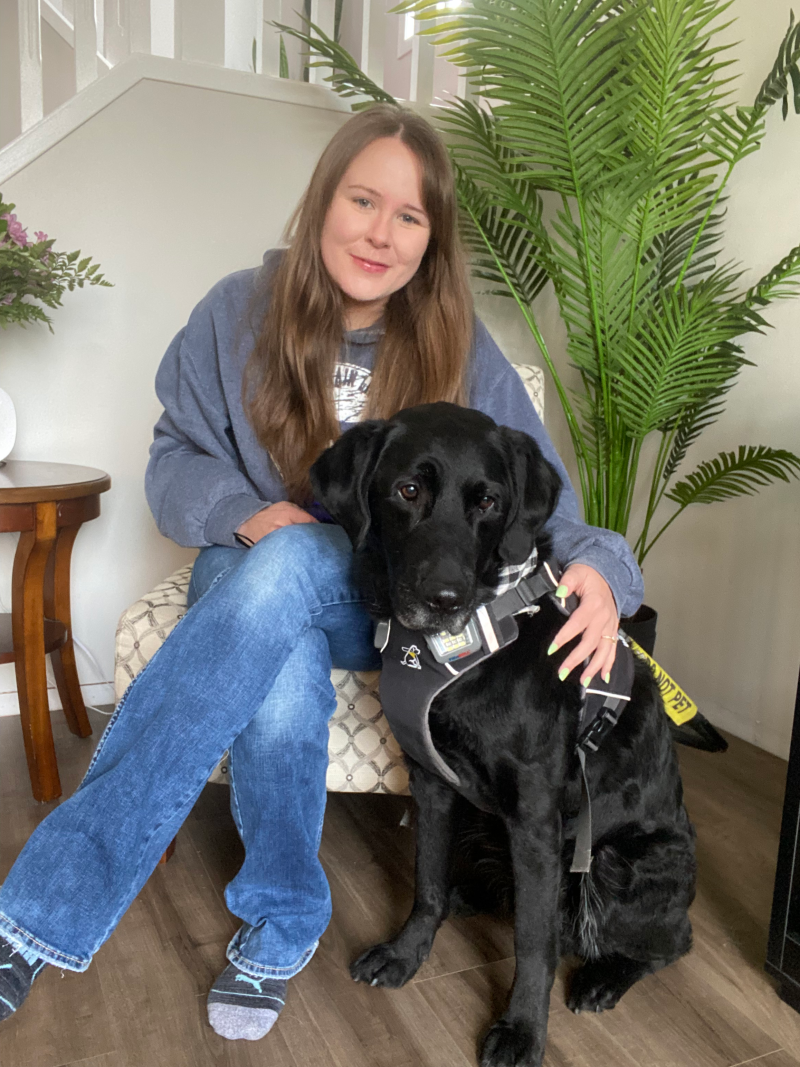 Taylor, a young woman with long brown hair, sits in a chair with her arm around her guide dog Wallace, a black lab wearing a CNIB harness, who sits beside her.