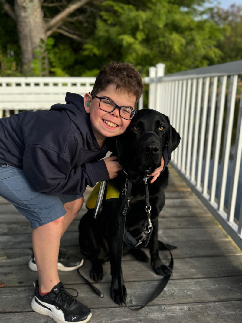Kaiden, a young boy, is crouching down and placing his arms around a black Labrador Retriever, Flinn.