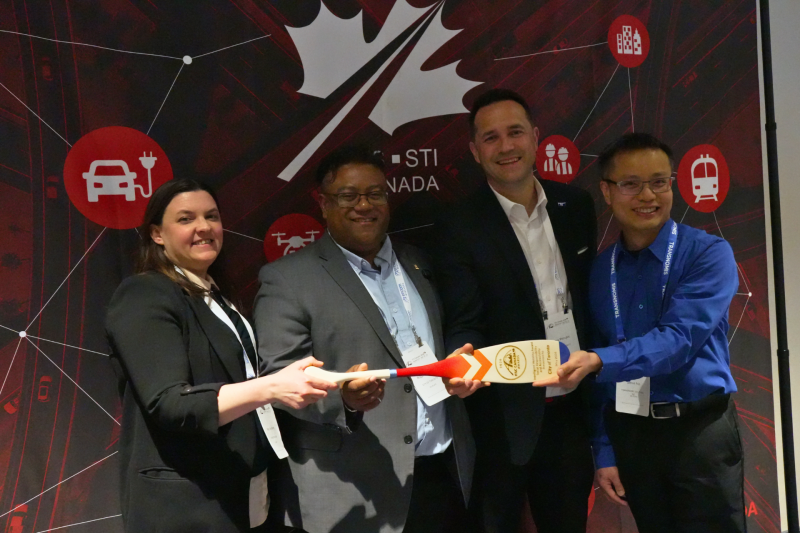 Four members of the Accessible Sidewalk Construction Zones for Blind and Low Vision Pedestrians pilot project team stand together at an event, smiling and holding their award, a wooden paddle engraved with the award information. Mary Ann, far left, holds one end of the paddle while the other team members gather around it.