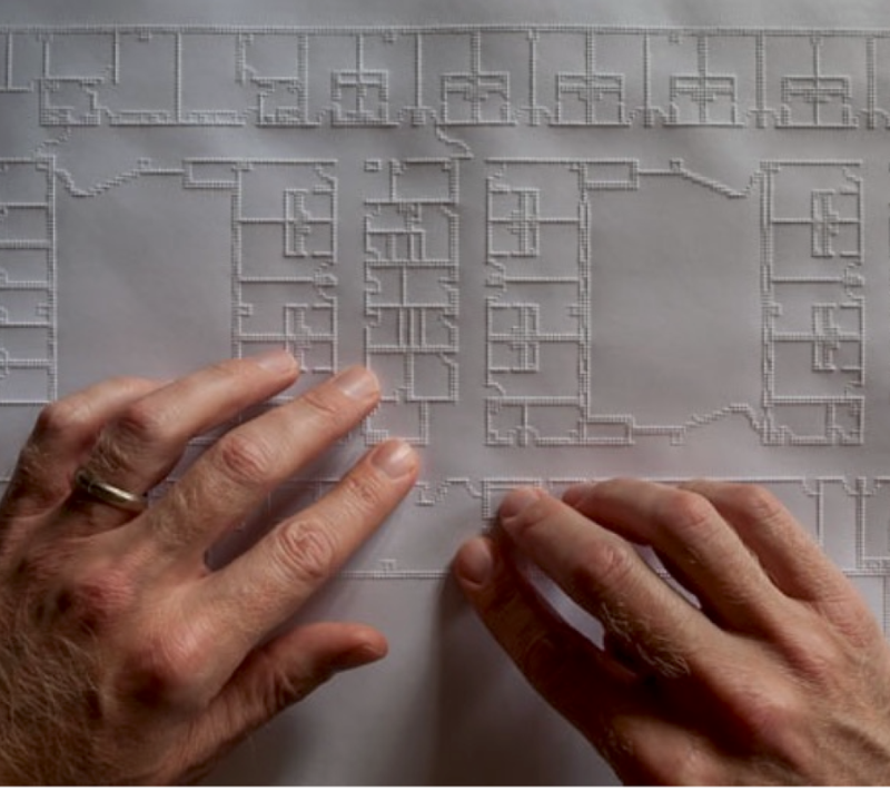 Two hands move across a large tactile architectural floor plan. Raised lines outline rooms, corridors, walls, and other features of the building layout.