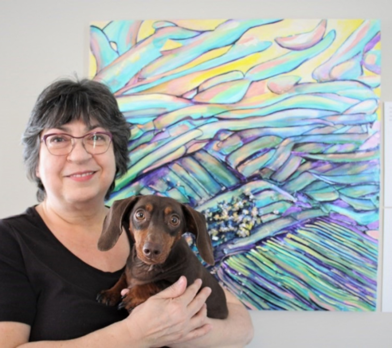Robyn Rennie poses for a photo in front of her colourful artwork next to a digital printed QR Code. She is holding a small, chocolate-brown dog.