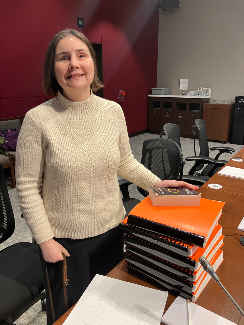 Jen Goulden smiles while standing at a desk in a Senate Committee meeting room. On the desk, she has her braille copy of the novel “The Fellowship of the Ring”, consisting of 6 thick, heavy volumes. On top of the pile sits a standard paperback version of the book, tiny in comparison. These props were used to demonstrate the high costs of shipping braille materials.