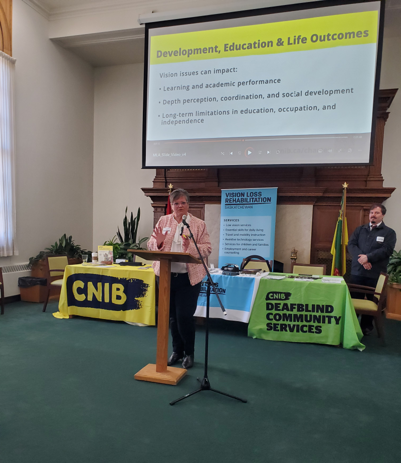 CNIB’s Chief Inclusion Officer, Christall Beaudry, stands at a podium and microphone to provide her remarks. Behind her are tables with the CNIB, DBCS, and VLR logos, along with a slideshow projected onto a large screen.