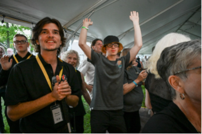 Chayse Zehr smiles and claps while standing among a crowd under a tent, as a person behind him raises both arms in the air.