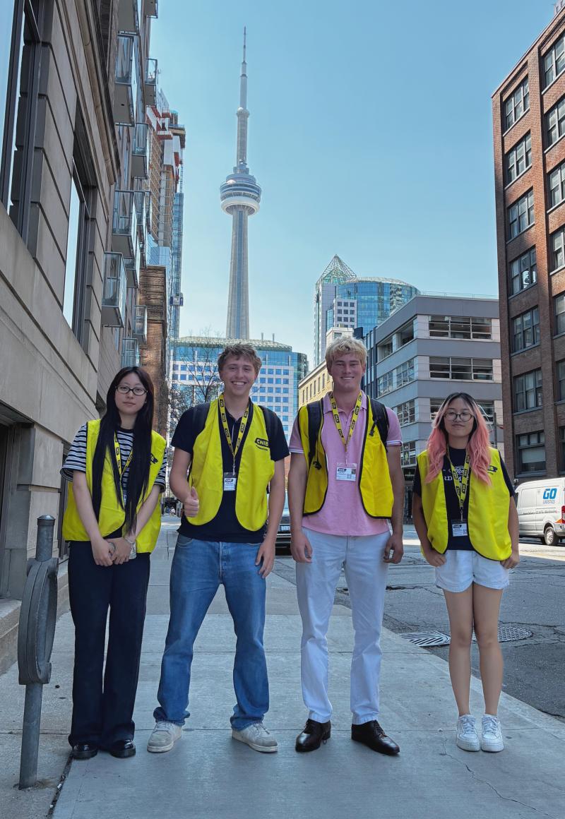Four canvassers wearing yellow CNIB vests and lanyards stand on a sidewalk with the CN Tower behind them.