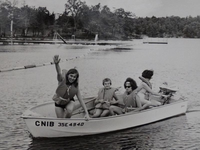 A black and white photo from the 1960s featuring 4 children with light skin wearing life jackets and sitting in a boat on a lake. The boat has the name CNIB painted on the side.