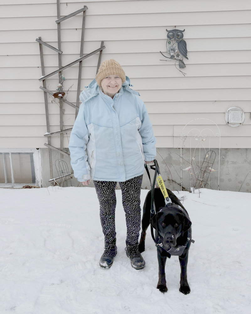 Barb stands in a snowy backyard with her black guide dog, Beacon, in harness at her side.