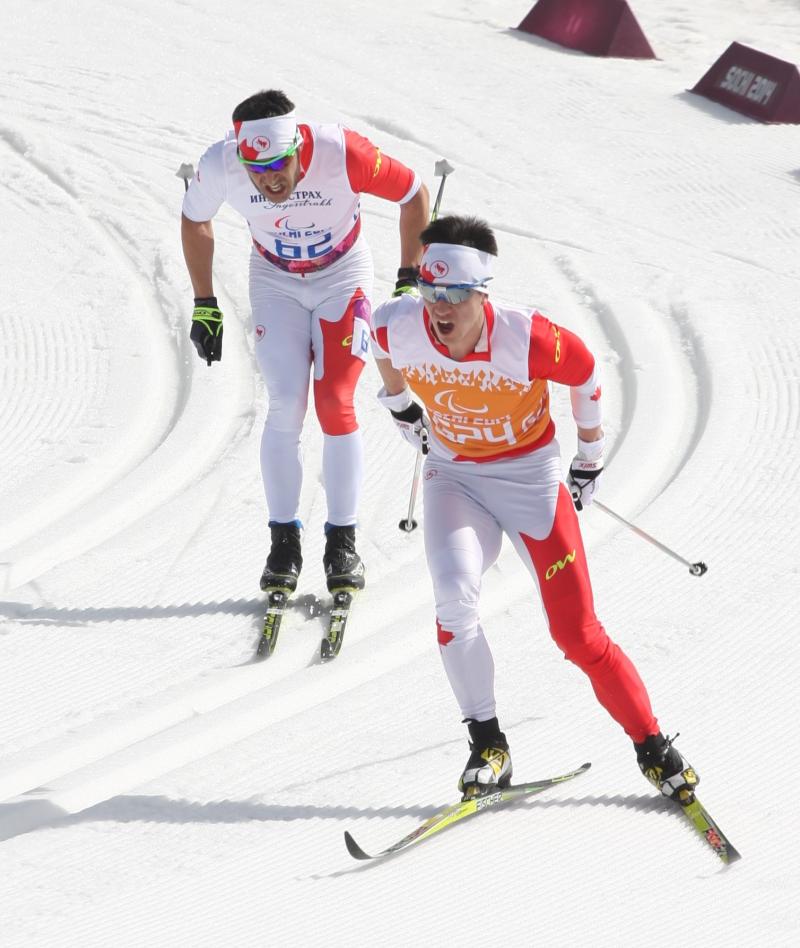 Brian McKeever and another skier in competition uniform ski down the slope.