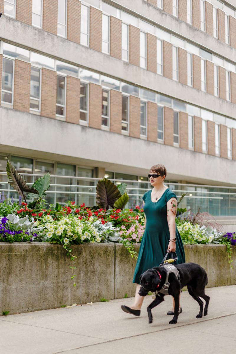 Denise and her guide dog Tara walking on the sidewalk near a large high-rise building.