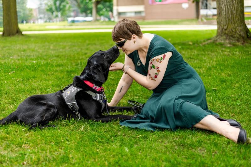 Denise and her guide dog Tara, a black Lab, sitting in the grass. Denise is gently holding Tara's face towards hers.