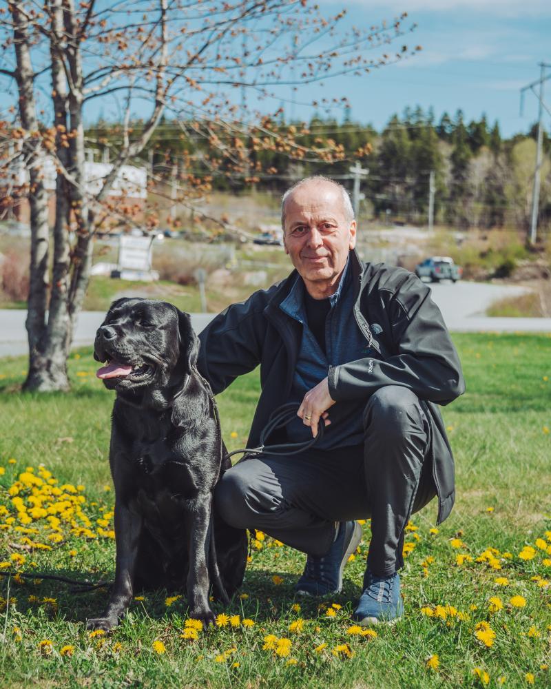 A man squats down beside a black Labrador retriever in a patch of grass. There is a tree with no leaves and a road behind them.
