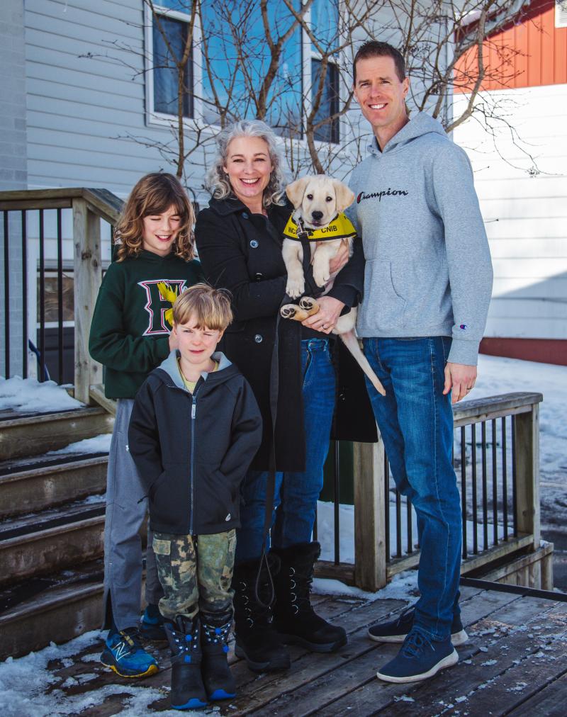 A family - a mother, father, and two young kids, pose on the front porch of their house. The mother, who is in the middle, is holding a golden retriever puppy. The puppy is wearing a yellow CNIB Guide Dog vest. There is snow on the ground.