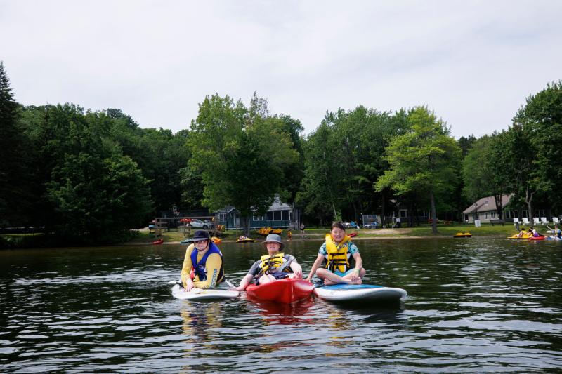 Three young people with light skin tones are on the lake. Two are sitting on paddleboards and one is sitting on a kayak.