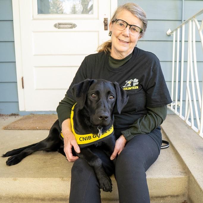 A woman wearing a CNIB Guide Dog Volunteer shirt sits on the front porch of a house with a CNIB guide dog overtop of her legs. The dog is a black Labrador retriever and is wearing a yellow CNIB Guide Dog vest.