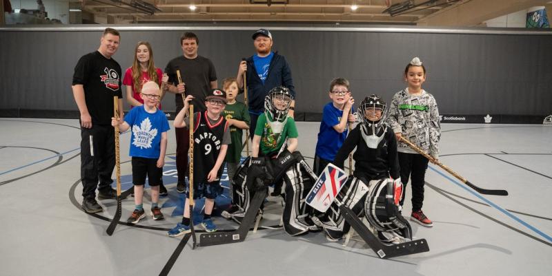 A group of children and Canadian Blind Hockey staff pose for their photo in a gymnasium. The participants have light to medium skin tones, and the children are wearing indoor hockey gear.