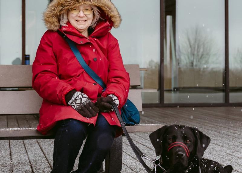 Cheri, who wears a hooded red parka, sits on a wooden bench with Sassy, her CNIB black Labrador guide dog, by her side on a snowy day.