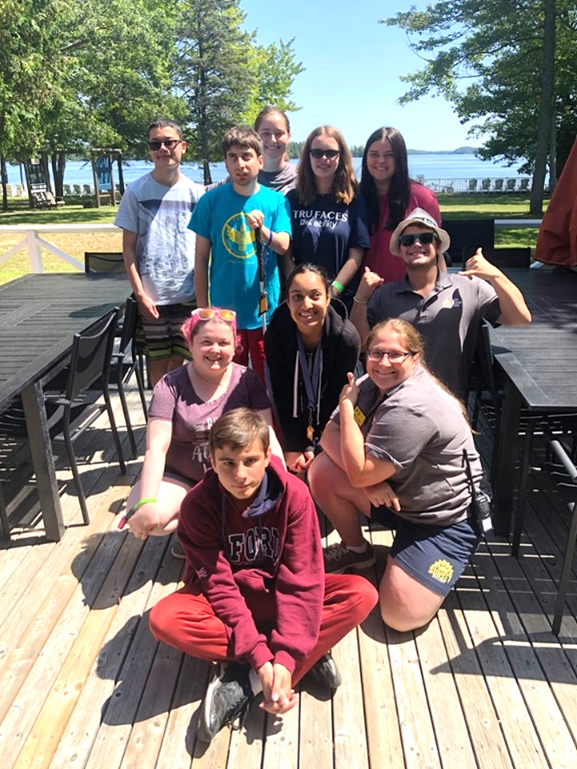 A group of young people with various skin tones smile for the camera in front of Lake Joseph during a Counsellor-in-Training Program.