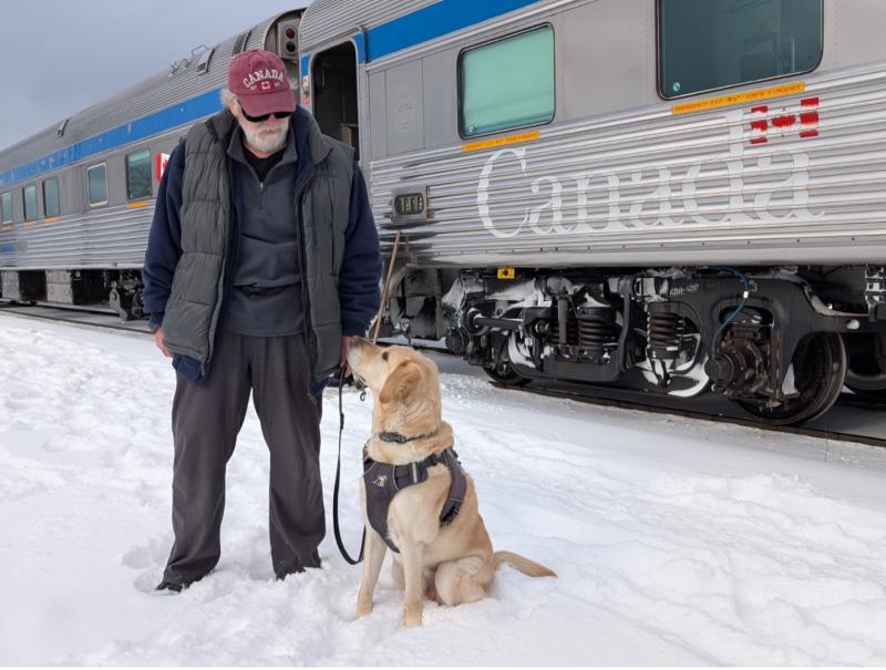 Dan stands in the snow beside his yellow Labrador guide dog, Sparky, who sits in harness and looks up at him. A VIA Rail passenger train is stopped on the tracks behind them.