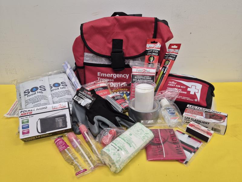 The contents of an emergency kit are shown spread out on a table. The items include an LED flashlight, emergency warming blanket, pocket radio, hand sanitizer, SOS food packets, and more.