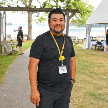 Eugene Chong smiling for the camera. Eugene is wearing a black shirt with black pants and has a yellow lanyard around his neck with a name tag attached. Lake Joseph is in the background of the picture.