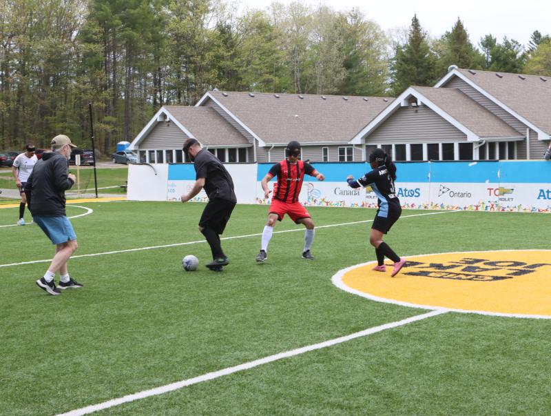 Blind soccer players, who have varying skin tones, wearing athletic clothing and eye shades, chase a soccer ball on the accessible soccer pitch at CNIB Lake Joe.