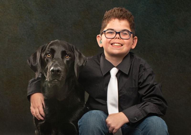 A child wearing glasses and a black shirt with a white tie sits beside a black dog, smiling.