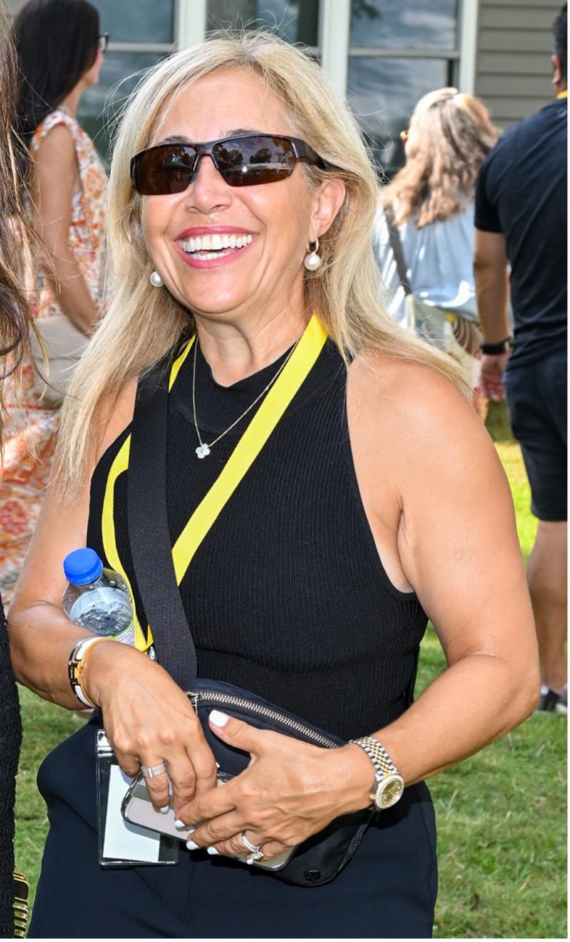 Frances is outdoors with a group of people milling about behind her. She is wearing dark glasses and smiling at the camera.