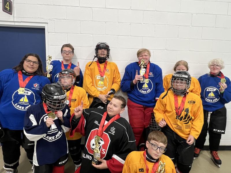 A group of children and young people - all of whom have light skin - pose in an arena wearing hockey equipment, including CNIB Lake Joe jerseys.
